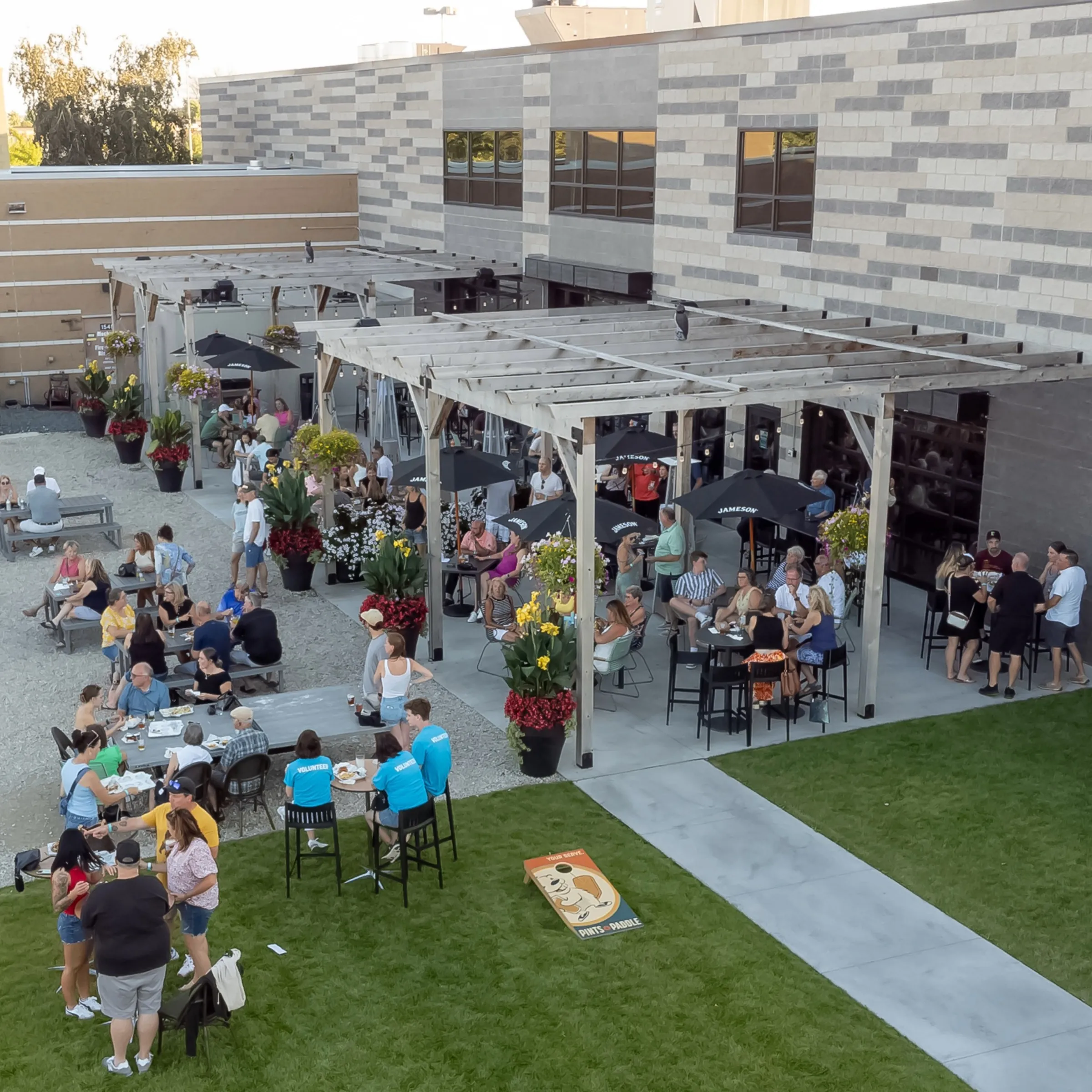 Golden-hour view of the patio with pergolas, umbrellas and a branded cornhole board on the lawn