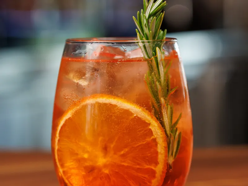 An Aperol spritz in a stemless wine glass with a large orange wheel and a tall rosemary sprig