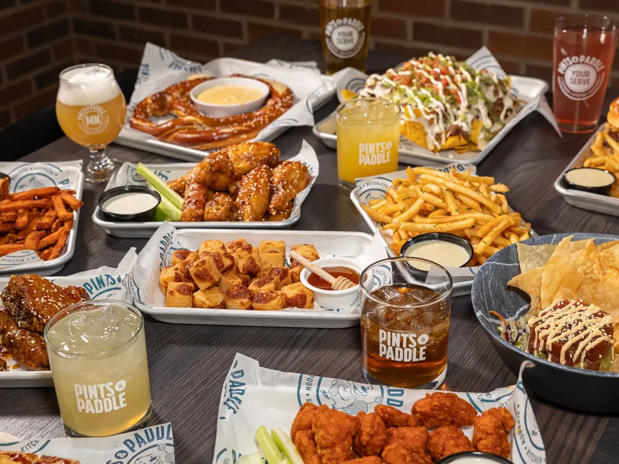 The full shareables table against the brick wall: wings, cheese curds, a giant pretzel, nachos, fries and drinks in logo glasses