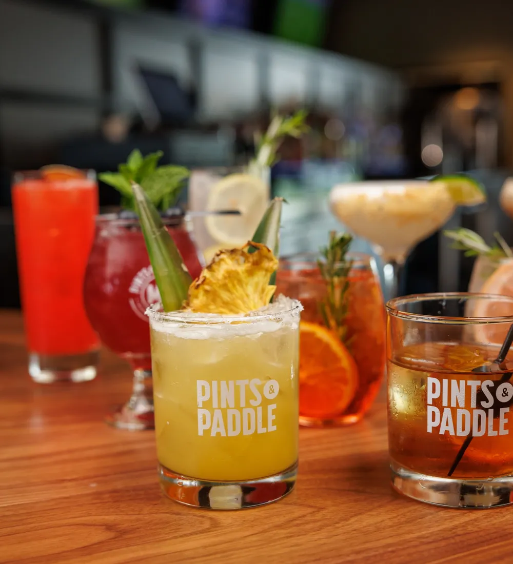 A charred pineapple margarita in a Pints & Paddle rocks glass front and center on the bar, an old fashioned beside it and a row of cocktails blurred behind