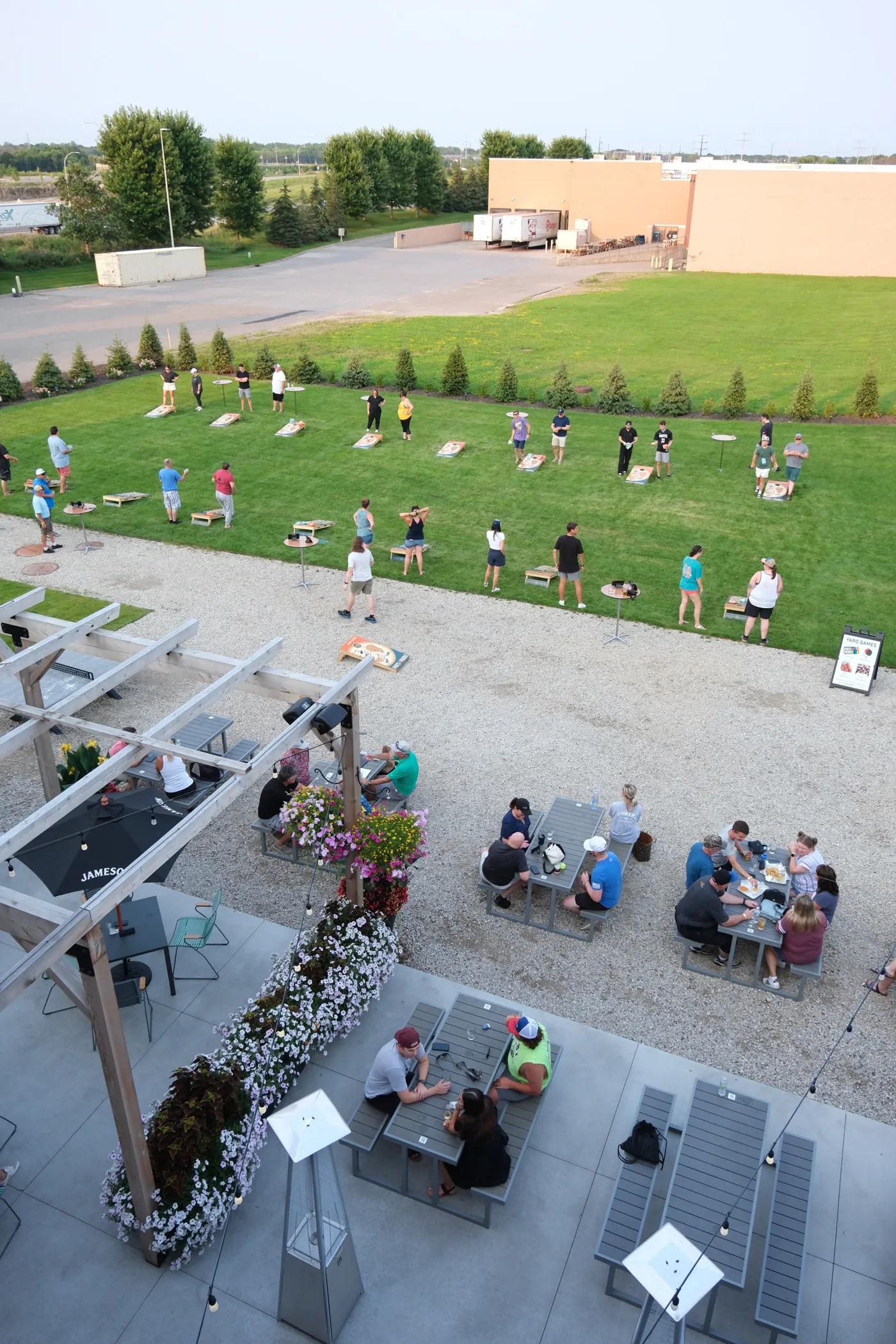 Rooftop view of the patio pergola and umbrellas with a bags league in progress on the lawn beyond, more than a dozen boards in play