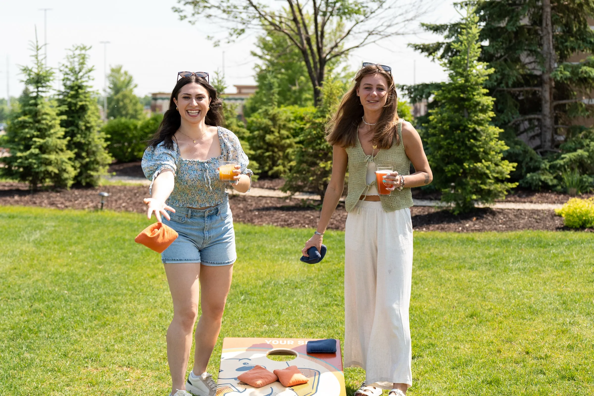Two friends playing bags on the lawn, one mid-toss with a drink in hand, a branded Pints & Paddle board in the foreground