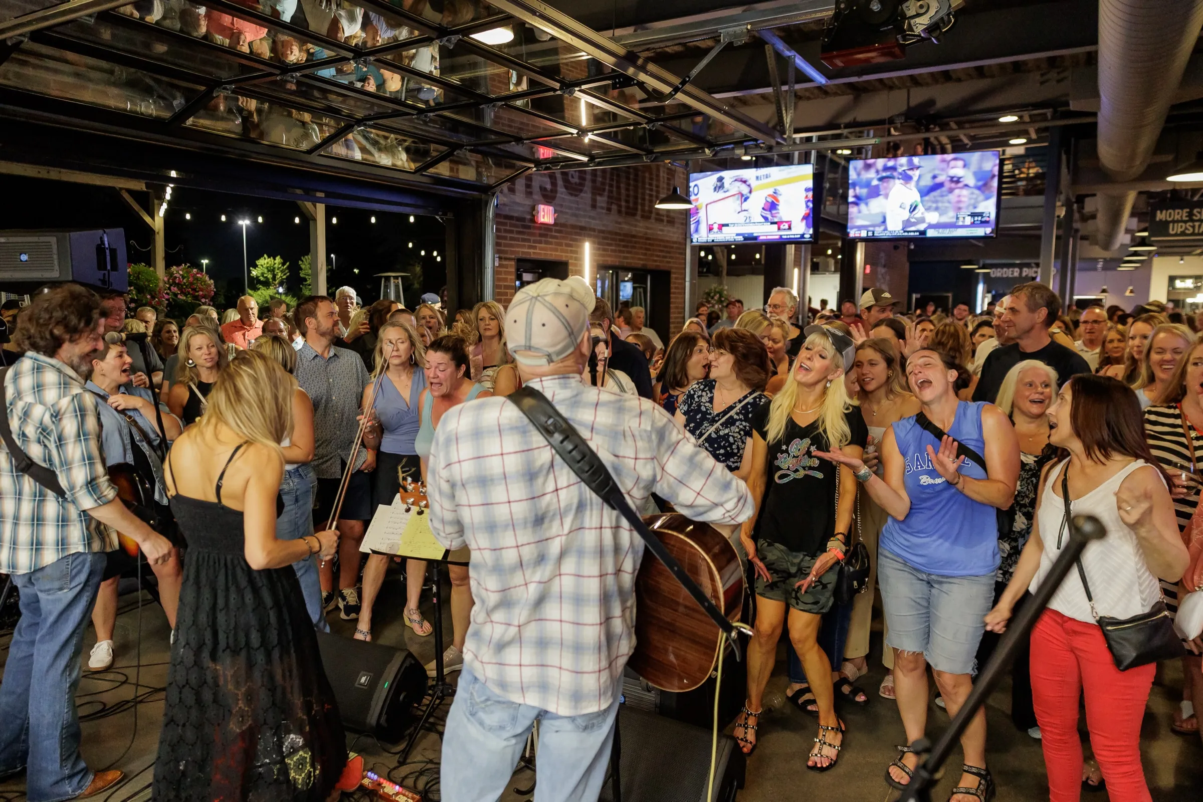 A band playing to a packed, singing crowd just inside the open garage doors at dusk, the lit patio behind them