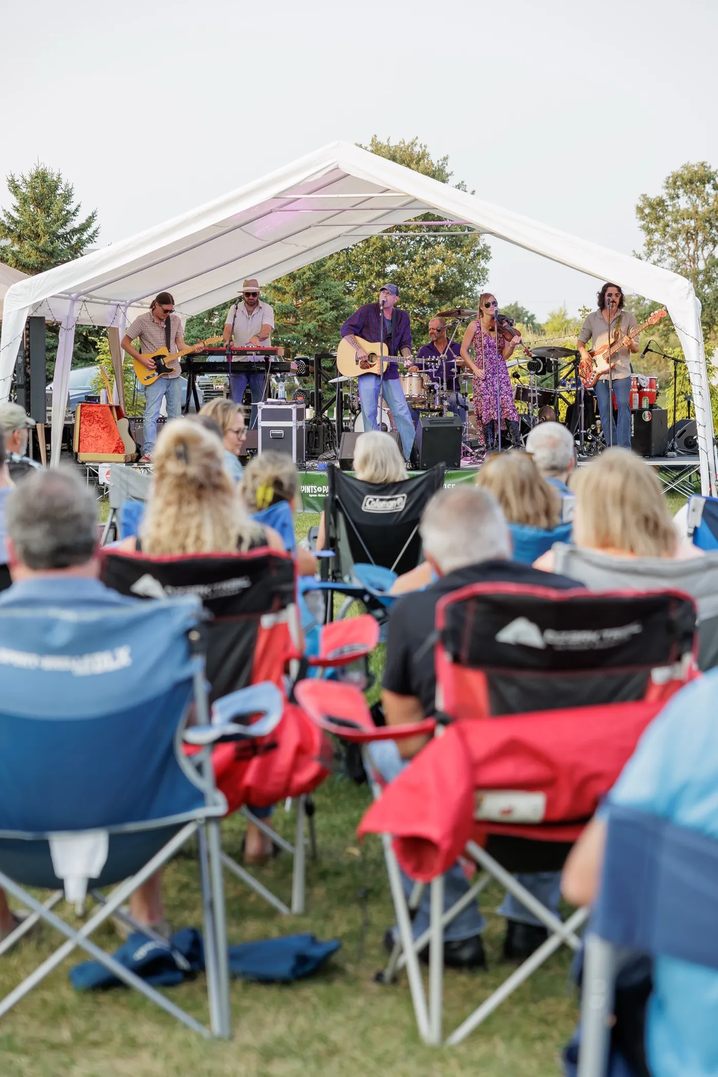 A band with guitars, keys, drums and a fiddle playing under a white tent on the lawn, the audience in camp chairs seen from behind
