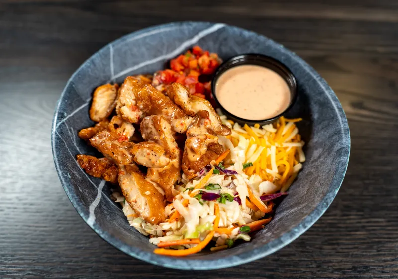A Bang Bang chicken bowl from above: crispy sauced chicken, slaw with carrot and purple cabbage, cheddar, pico and a ramekin of bang bang sauce