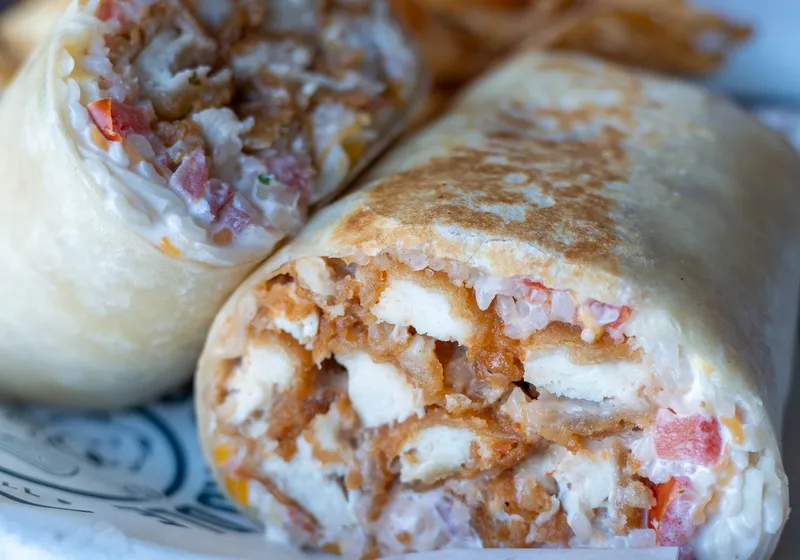 A toasted Bang! Bang! Chicken Wrap cut in half, showing crispy chicken, rice, pico and cheese on Pints & Paddle paper, with kettle chips behind it