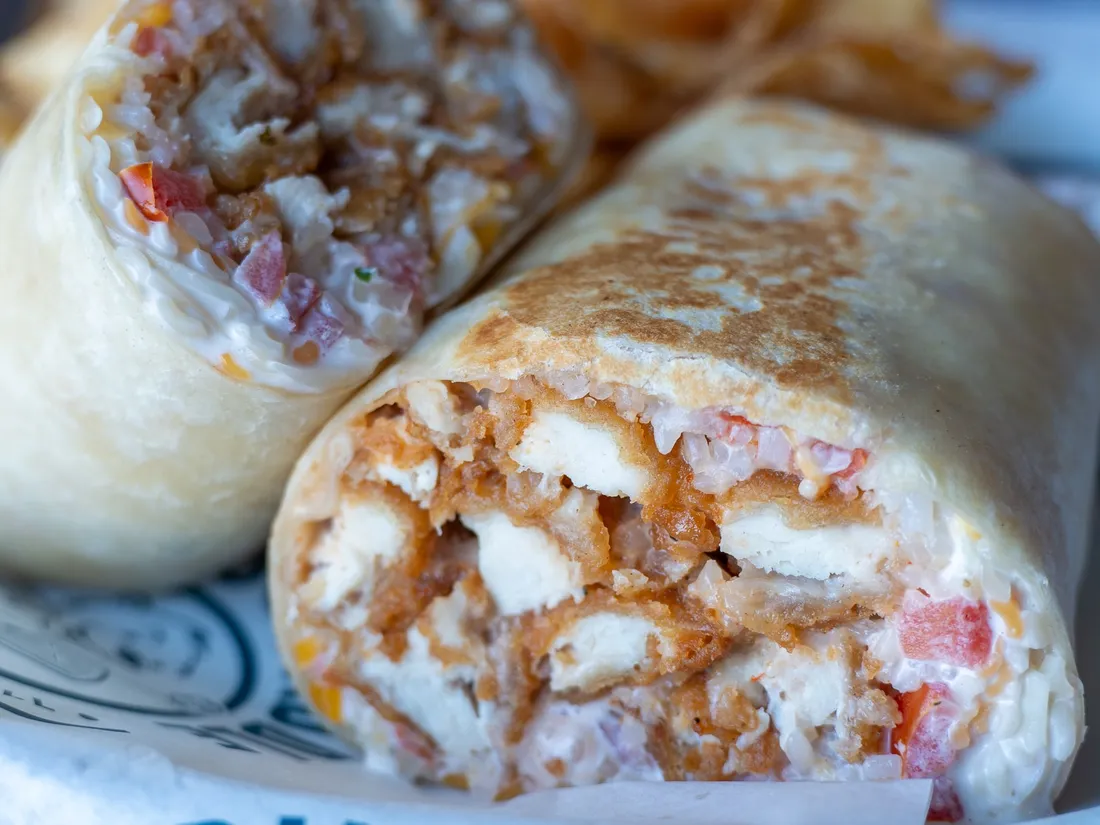 A toasted Bang! Bang! Chicken Wrap cut in half, showing crispy chicken, rice, pico and cheese on Pints & Paddle paper, with kettle chips behind it