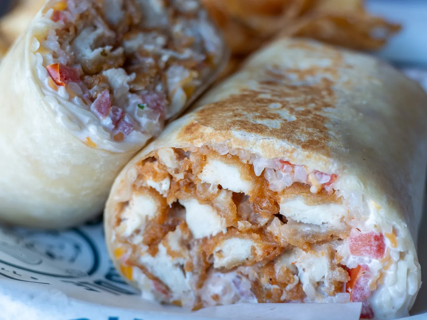 A toasted Bang! Bang! Chicken Wrap cut in half, showing crispy chicken, rice, pico and cheese on Pints & Paddle paper, with kettle chips behind it