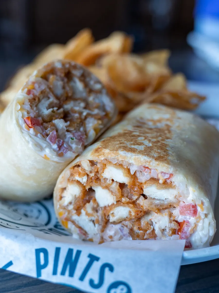 A toasted Bang! Bang! Chicken Wrap cut in half, showing crispy chicken, rice, pico and cheese on Pints & Paddle paper, with kettle chips behind it