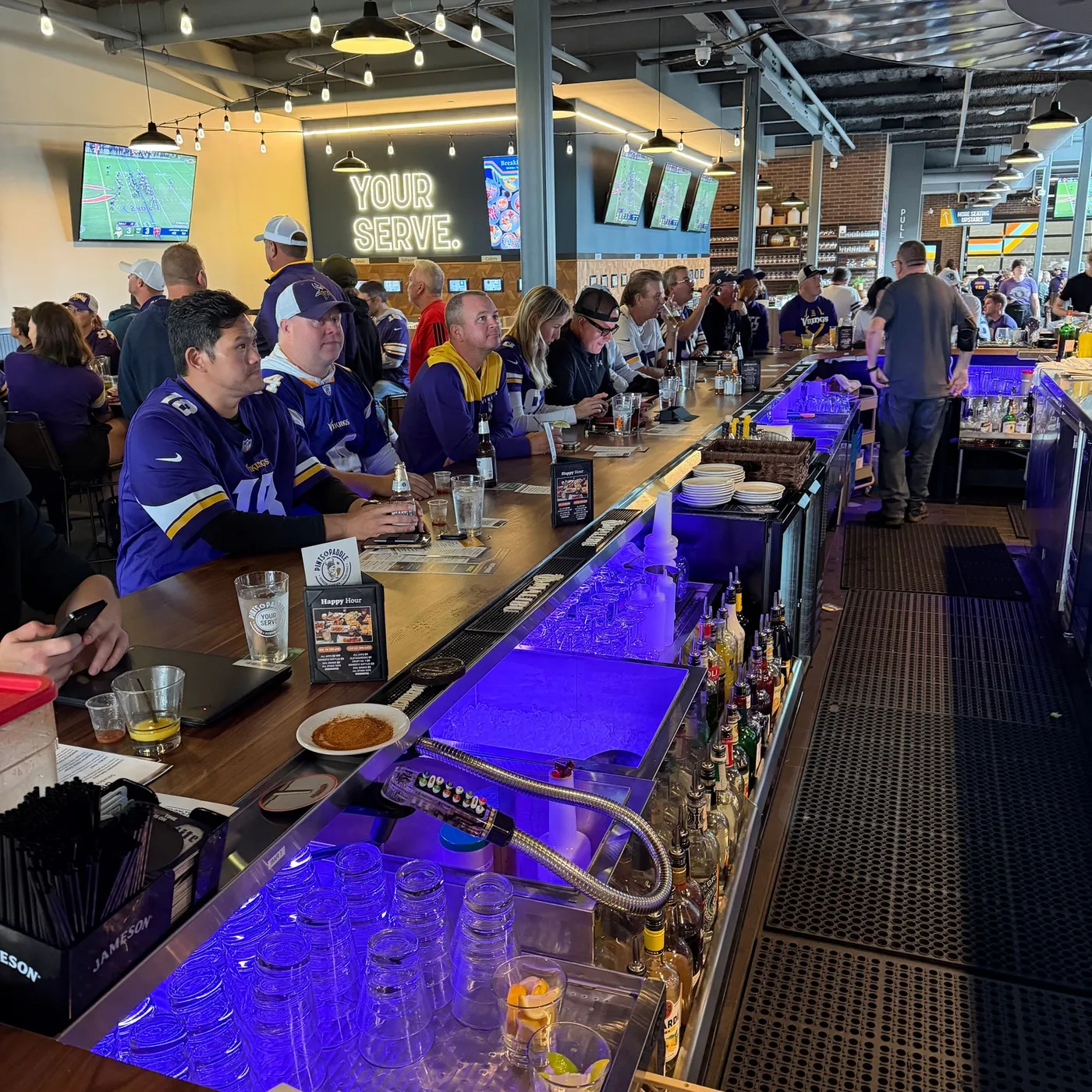 Fans in Vikings jerseys lined up along the bar under the glowing YOUR SERVE sign, the game on the TVs and the ice wells lit purple