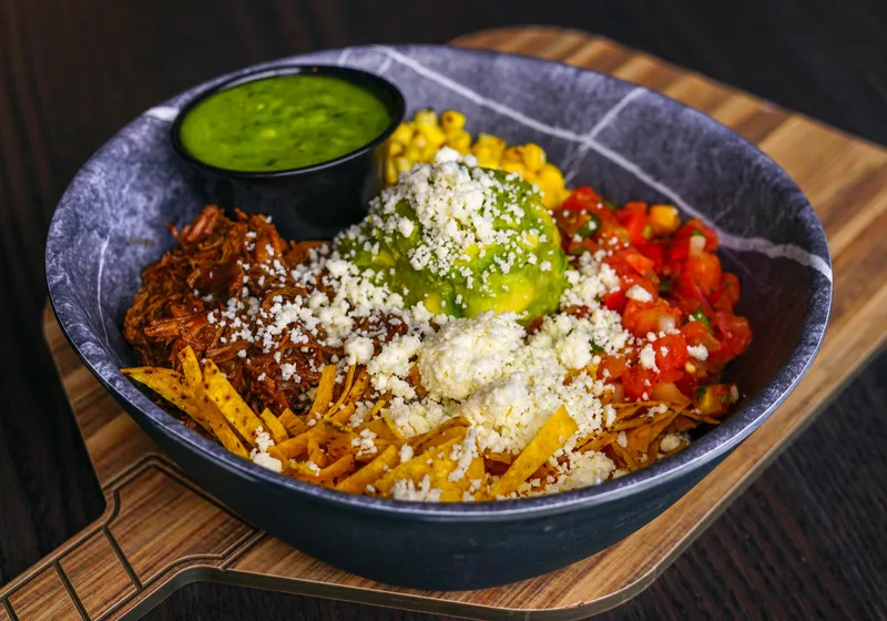 A barbacoa bowl with shredded beef, guacamole topped with cotija, tortilla strips, corn and pico, with a cup of salsa verde, on a paddle-shaped board