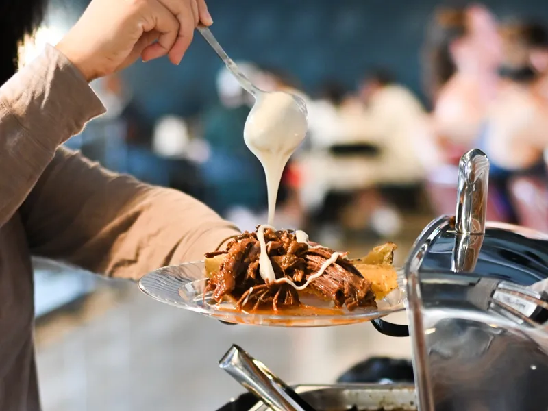 A guest drizzles a spoonful of queso over a plate of shredded barbacoa at the buffet, a chafing dish lid at right and other diners blurred behind
