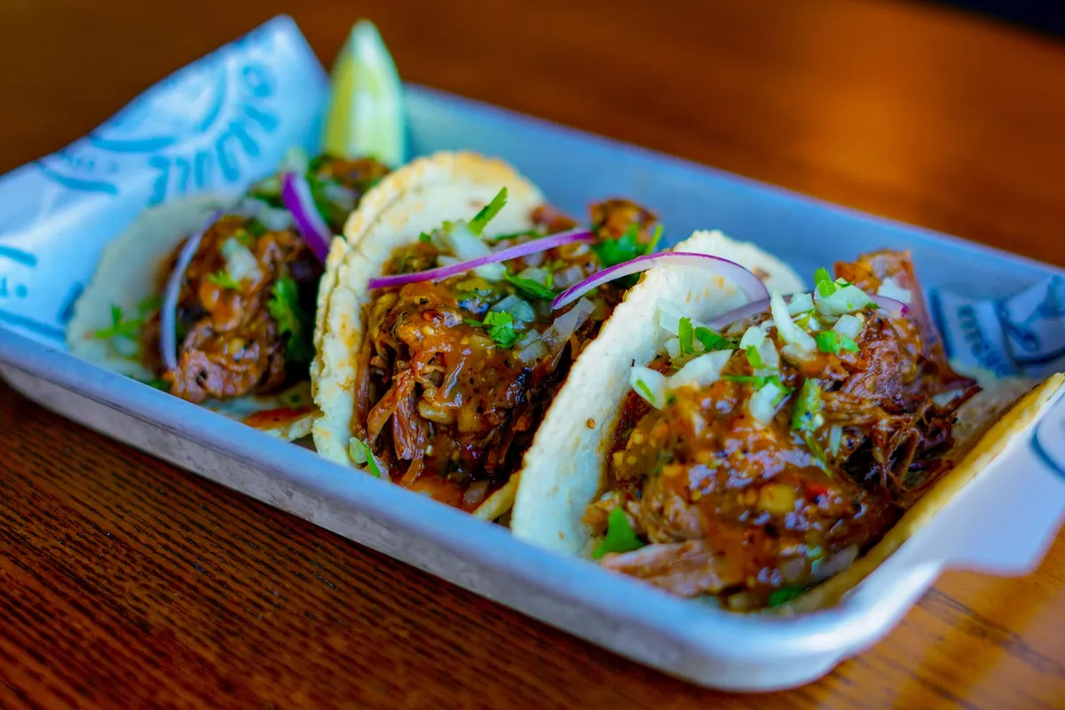 Three barbacoa street tacos with red onion and cilantro in a metal tray, lime wedge behind