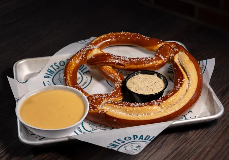 A giant salted Bavarian pretzel on a branded-paper tray with a bowl of cheese dip and a cup of grain mustard