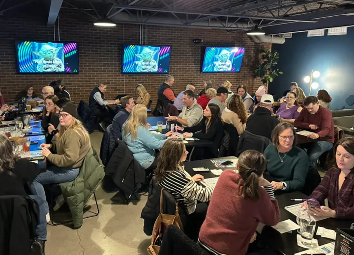 A full mezzanine crowd at tables with bingo cards and pens, three screens lit over the tap wall