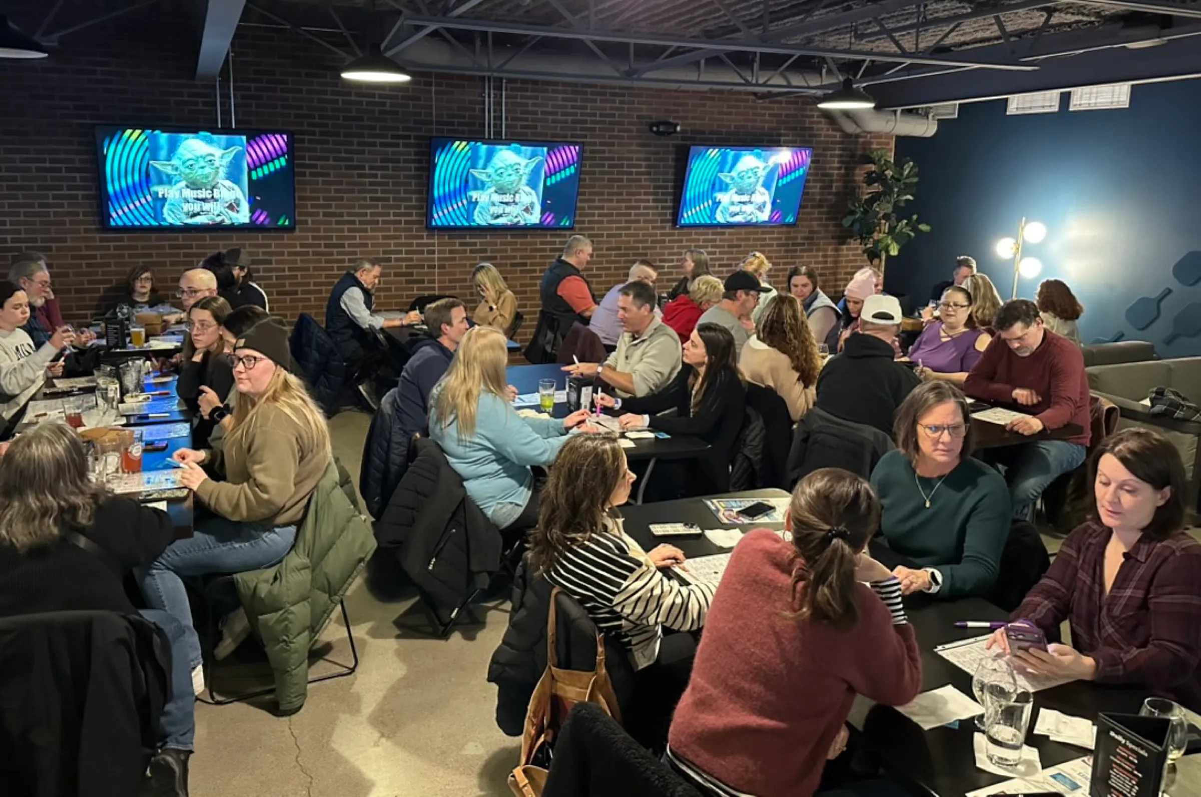 A full mezzanine crowd seated at long tables with bingo cards and pens, three screens lit over the tap wall