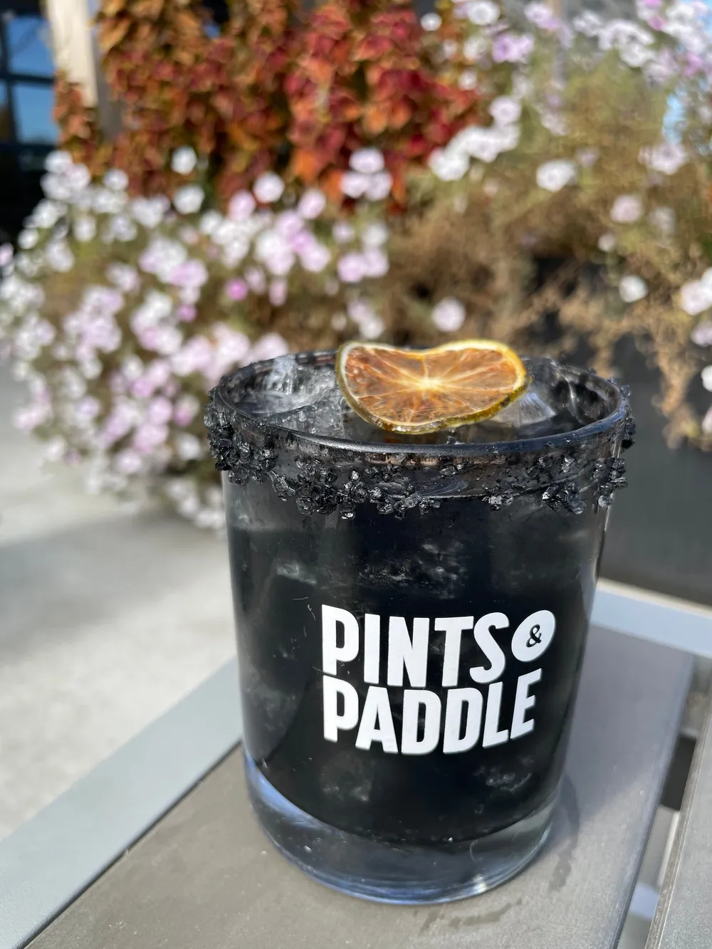 The Black Magic Margarita, jet black over ice with a black salt rim and a dried lime wheel in a Pints & Paddle rocks glass, patio flowers behind