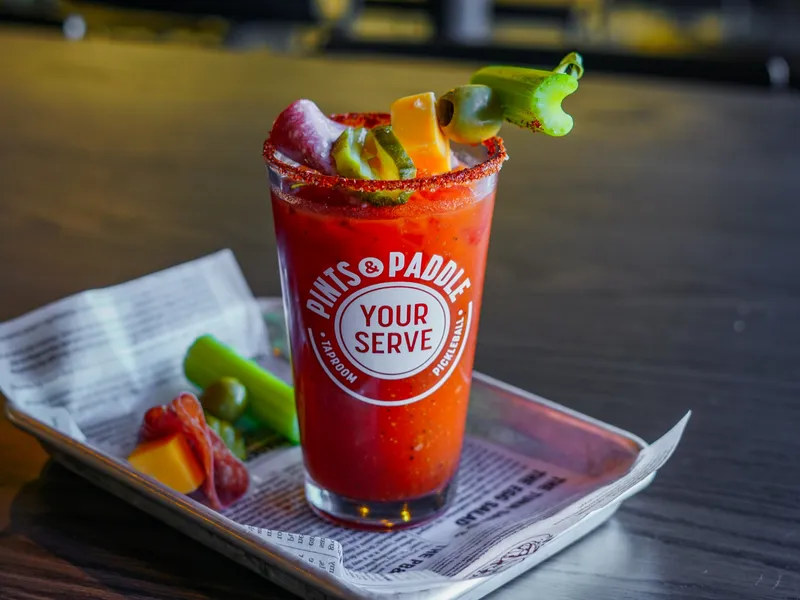 A Bloody Mary in a branded pint glass with a spiced rim and a skewer of celery, cheese, pickle, olive and salami