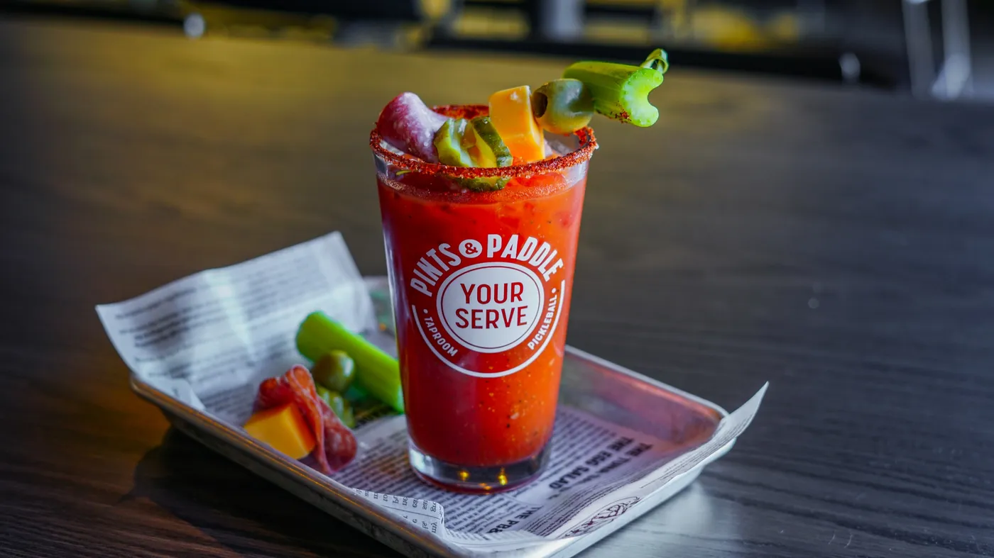 A Bloody Mary in a branded pint glass with a spiced rim and a skewer of celery, cheese, pickle, olive and salami, with an extra garnish plate beside it