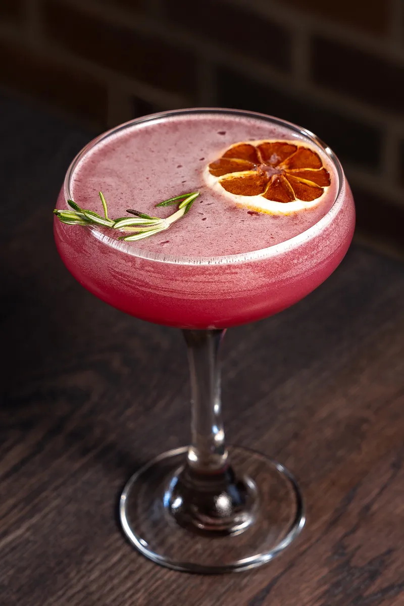 A magenta blueberry lemon drop in a coupe with a dried citrus wheel and a rosemary sprig against a brick wall