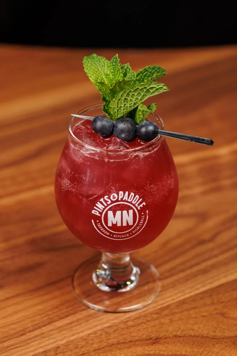 A deep red-purple blueberry mint mocktail in a Pints & Paddle MN tulip glass with a mint bouquet and three blueberries on a pick