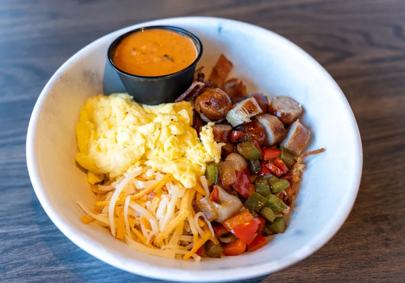 A breakfast bowl of scrambled eggs, hash browns, sausage, peppers, onions and cheese with a cup of sauce