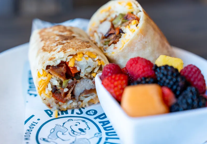 A griddled breakfast wrap cut in half beside a cup of fresh berries, pineapple and melon