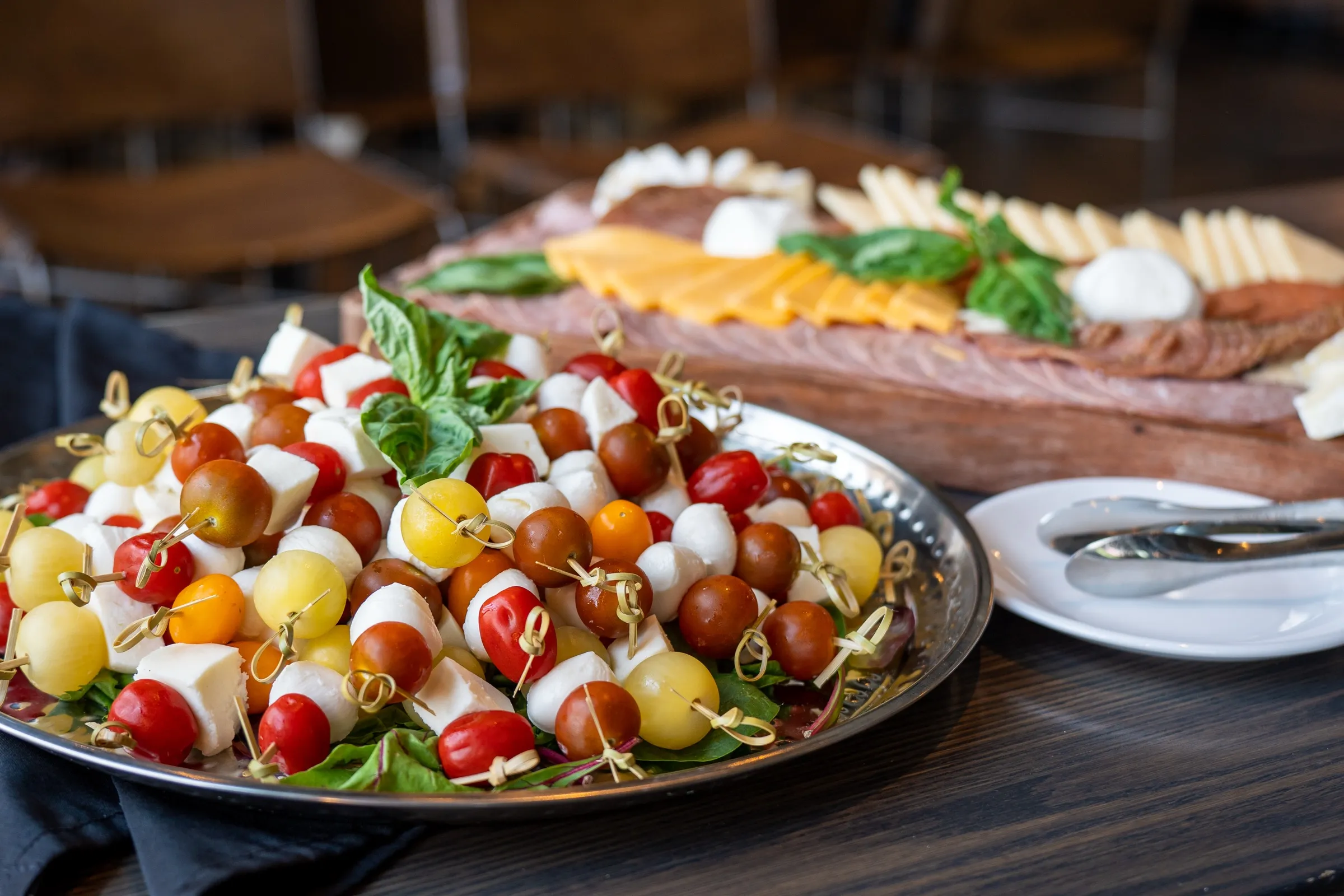 A silver platter of caprese skewers in front of a wooden charcuterie board of cheddar, salami and brie