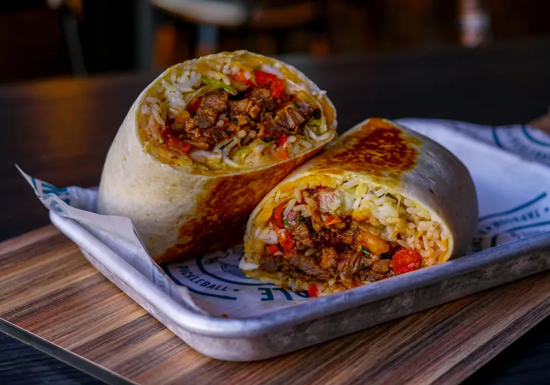 A carne asada wrap cut in half showing steak, rice, lettuce and pico, stacked on branded parchment in a metal tray