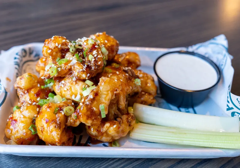 A pile of glazed crispy cauliflower bites with sesame seeds and scallions, celery sticks and a ramekin of ranch on a branded tray