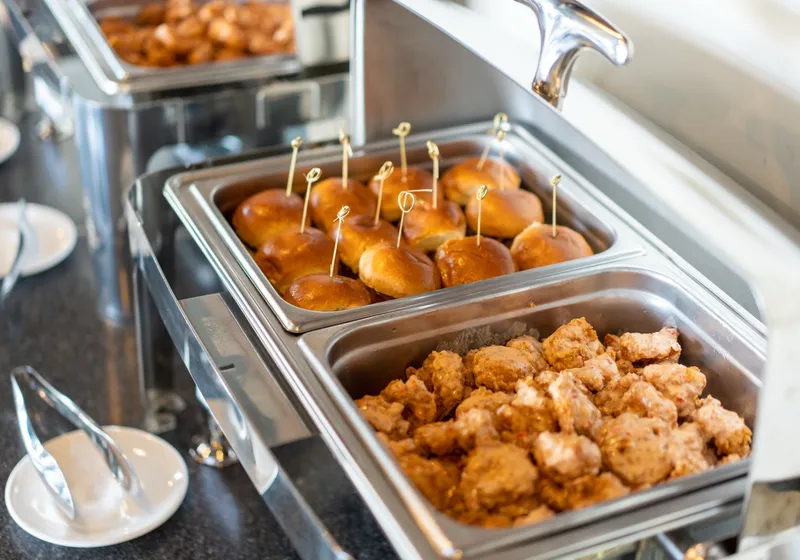 Stainless chafing dishes of brioche slider buns and boneless wings with small plates and tongs lined up on a dark counter