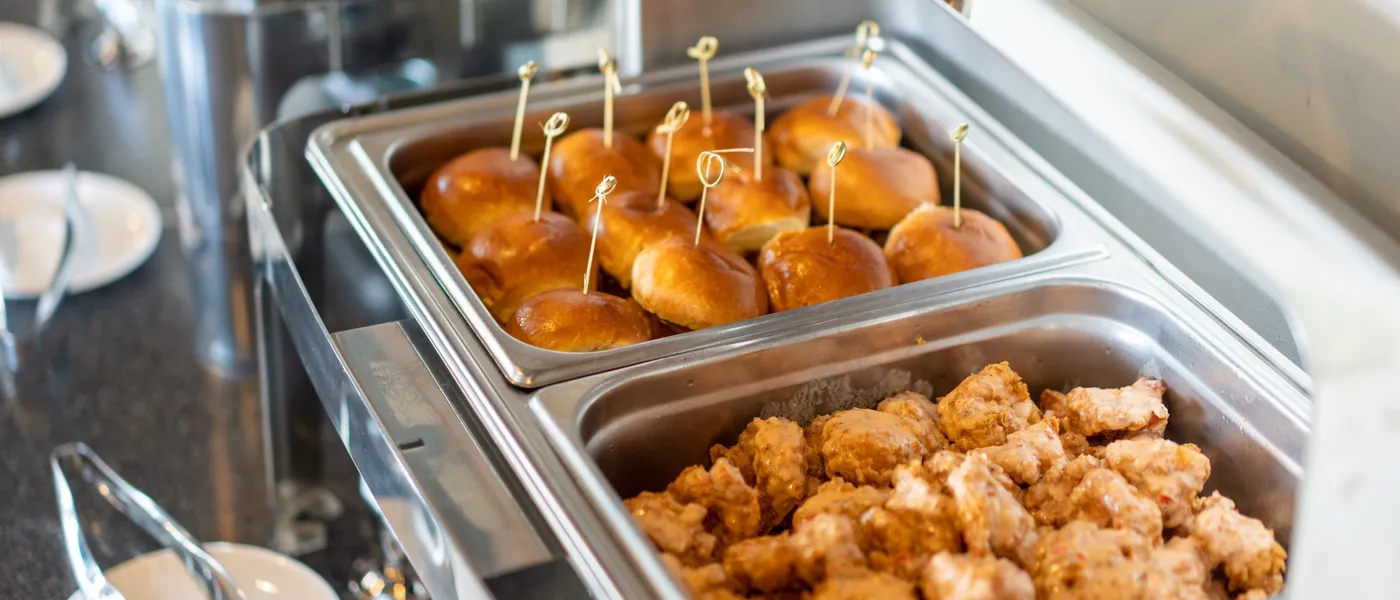 A buffet line of stainless chafing dishes: brioche slider buns with skewer picks beside a pan of boneless wings, small plates and tongs lined up on the dark counter