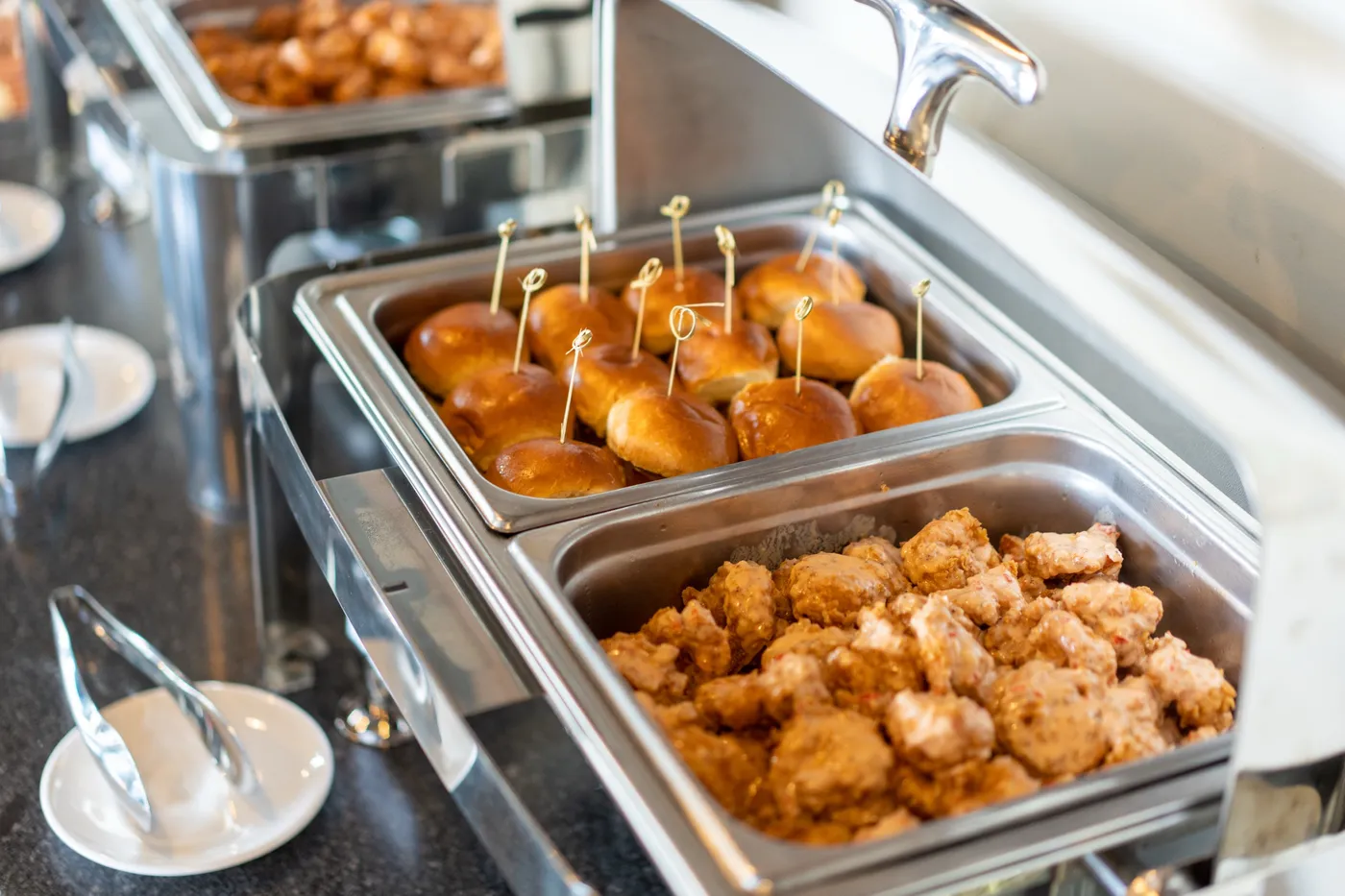 Stainless chafing dishes of brioche slider buns and boneless wings with small plates and tongs lined up on a dark counter