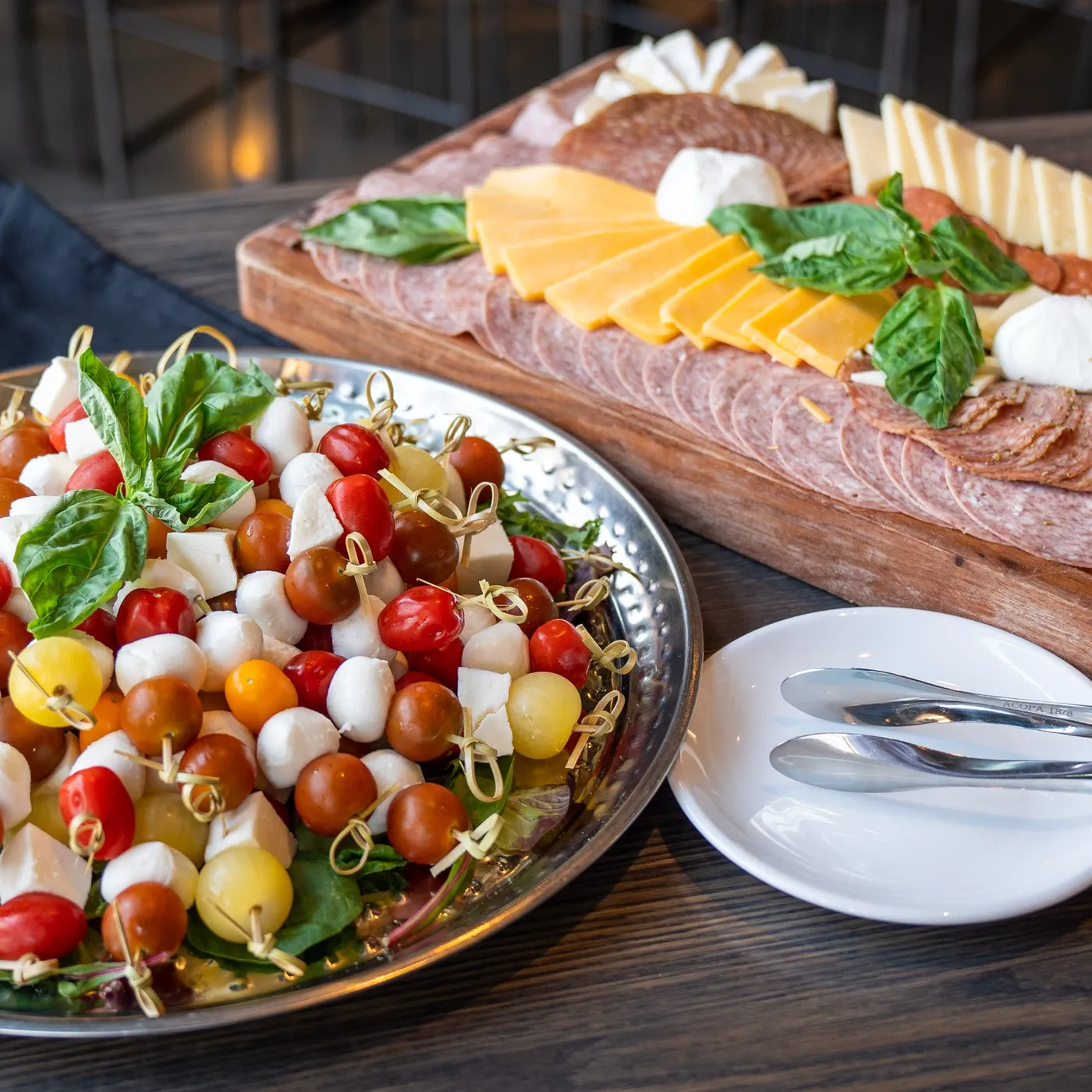 A silver platter of caprese skewers beside a wooden charcuterie board of cheddar, salami and brie