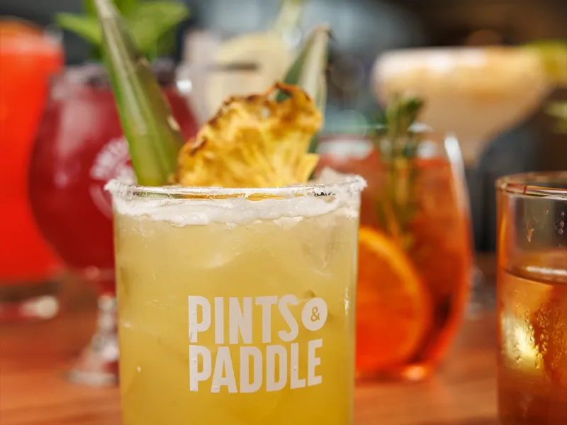 A charred pineapple margarita in a salt-rimmed Pints & Paddle rocks glass with a charred pineapple wedge and fronds