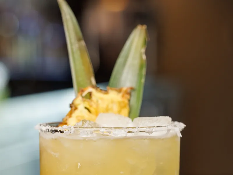 A charred pineapple cocktail in a salt-rimmed Pints & Paddle rocks glass with a pineapple wedge and leaves against the lit back bar