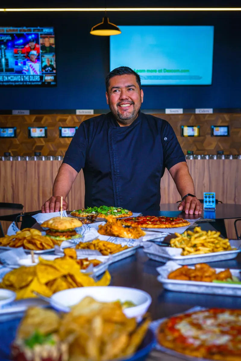Executive Chef Noé Lara smiling behind a spread of pizzas, burgers and nachos in the taproom