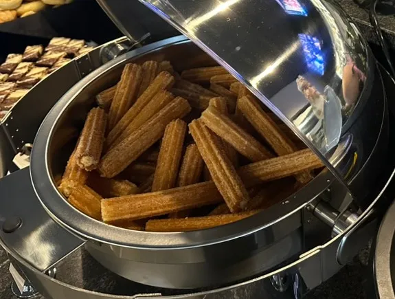 Cinnamon-sugar churros heaped in a round stainless chafer with the dome lid rolled back, dessert trays behind