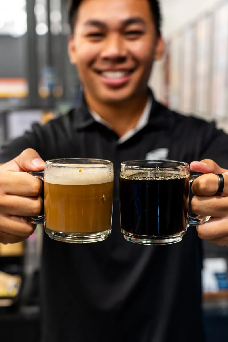 A smiling team member in a black polo holds out two glass mugs, one of coffee with a foamy top and one of black coffee