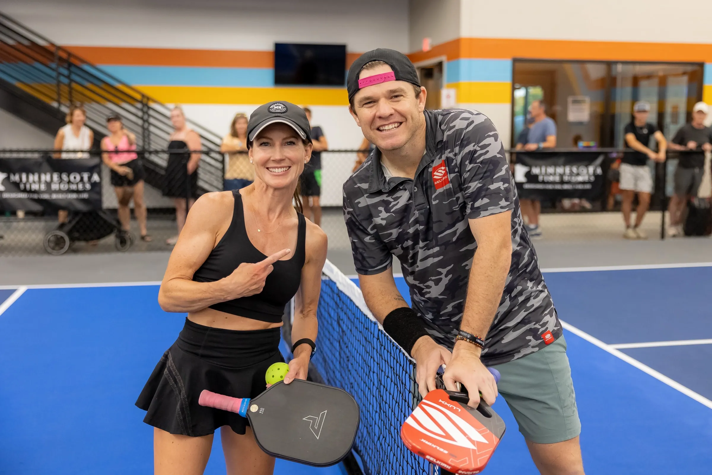 A smiling mixed doubles pair posing at the net with paddles and a ball