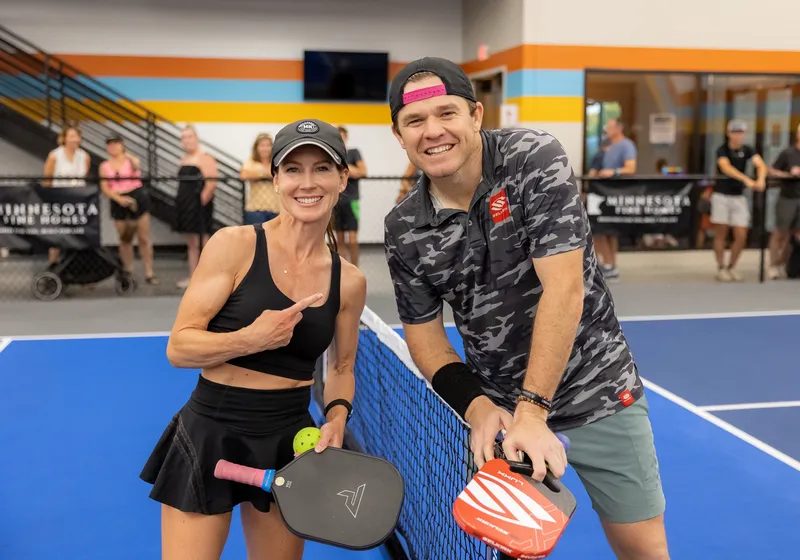 A smiling mixed doubles pair pose at the net with paddles and a ball