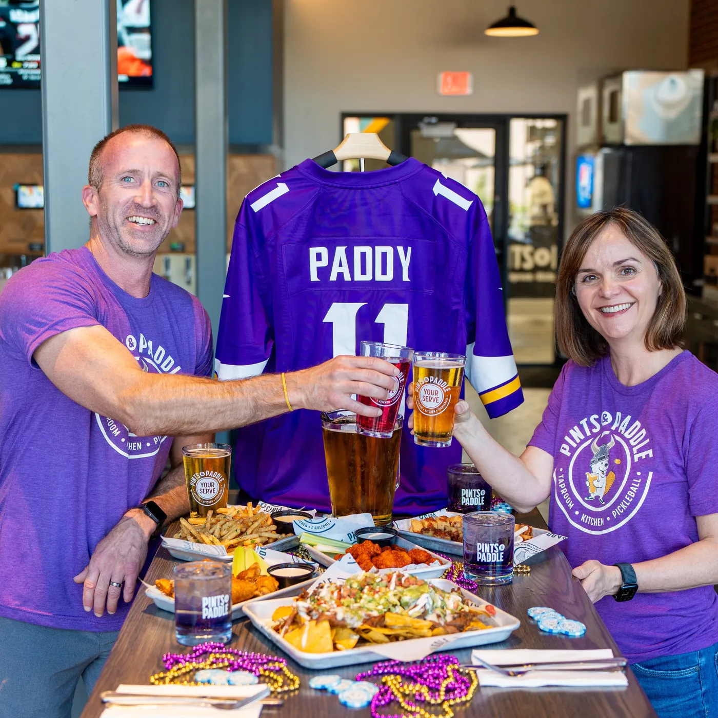 A man and a woman in purple Pints & Paddle tees clink pint glasses over a table of nachos, wings and fries, a purple Paddy 11 jersey hanging between them