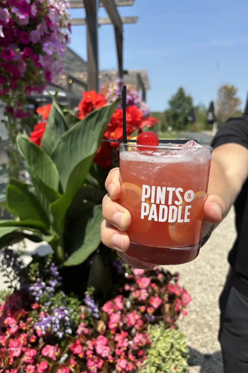 A hand holds up a Pints & Paddle rocks glass of pink-red Court No. 9 with lemon wheels and a cherry against the patio flowers