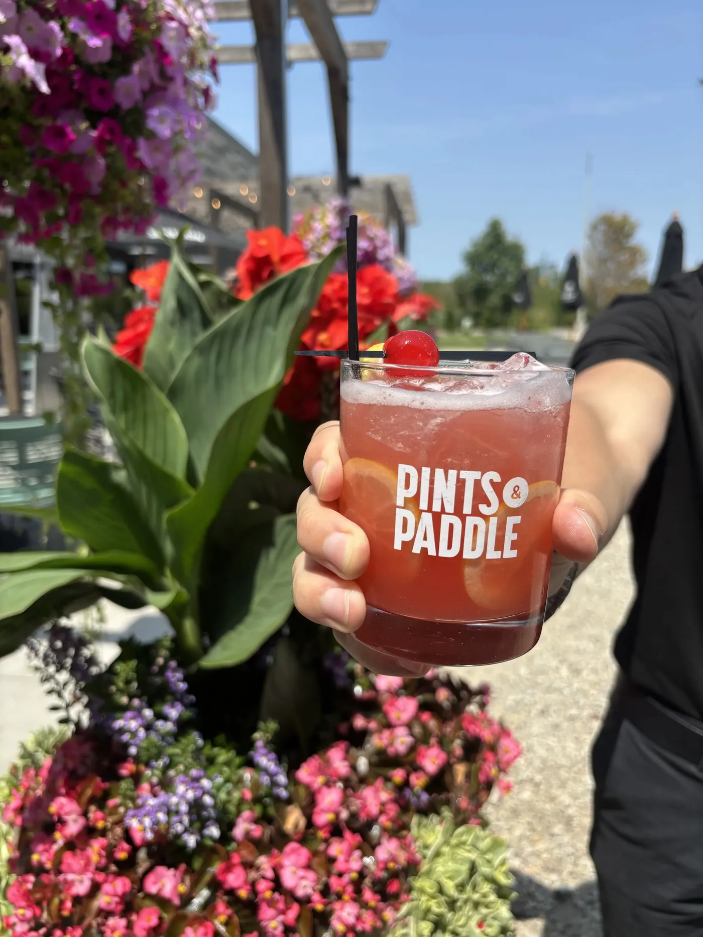 A guest holds up a Pints & Paddle rocks glass of Court No. 9, pink and bright with lemon wheels and a cherry, against the patio flowers