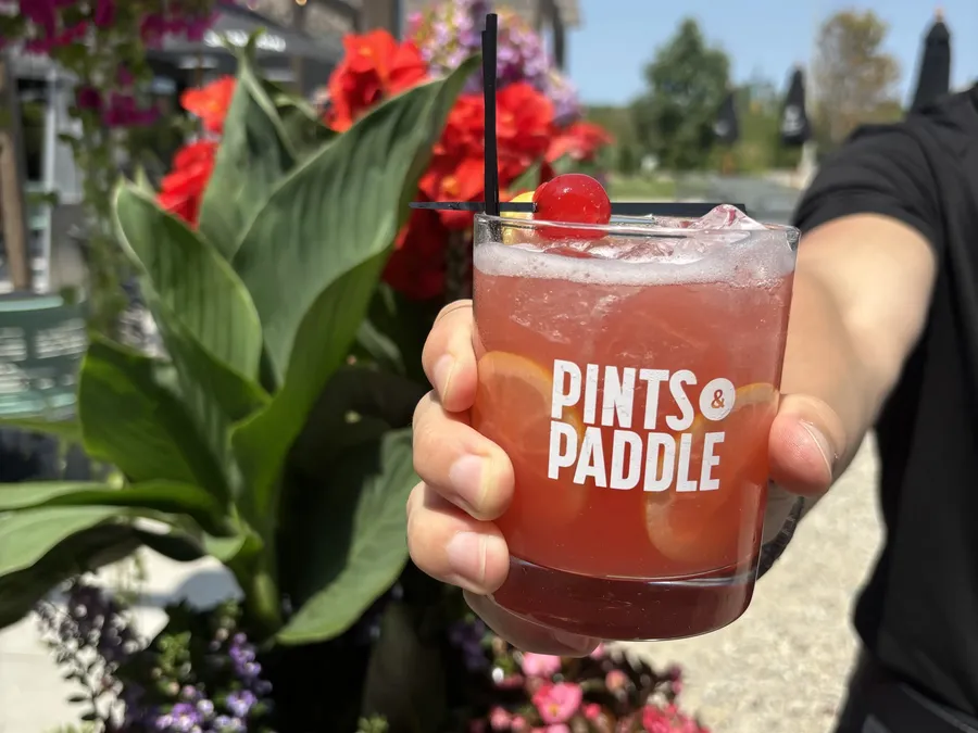 A hand holds up a Pints & Paddle rocks glass of pink-red Court No. 9 with lemon wheels and a cherry against patio flowers