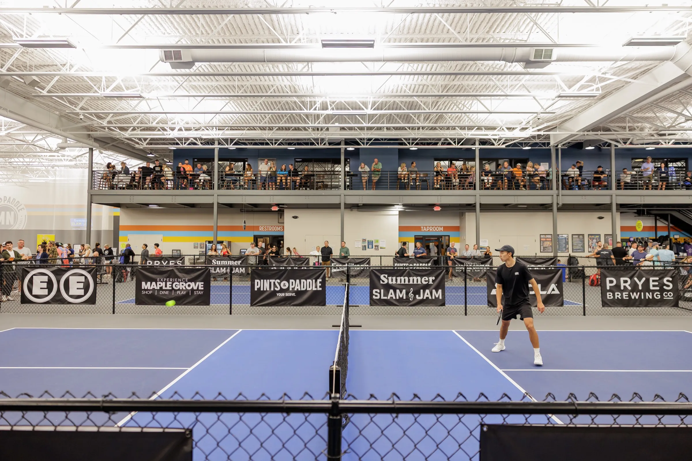 Wide view of the indoor courts with a player waiting at the net, sponsor banners along the fences and a full mezzanine above