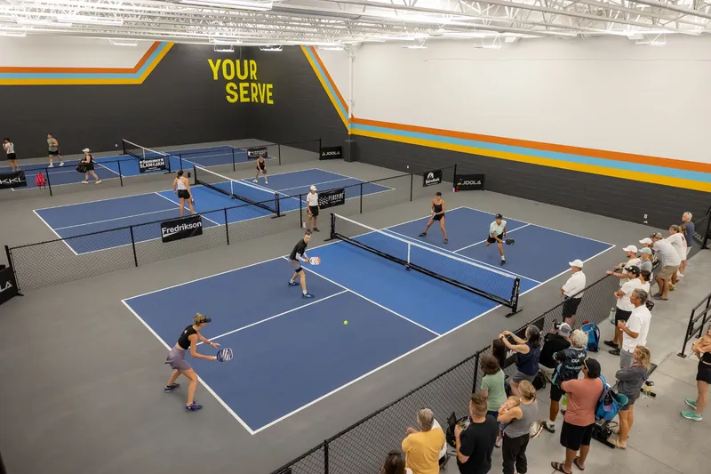 View from the mezzanine over three blue courts with doubles matches in play under the yellow Your Serve wall, spectators along the fence