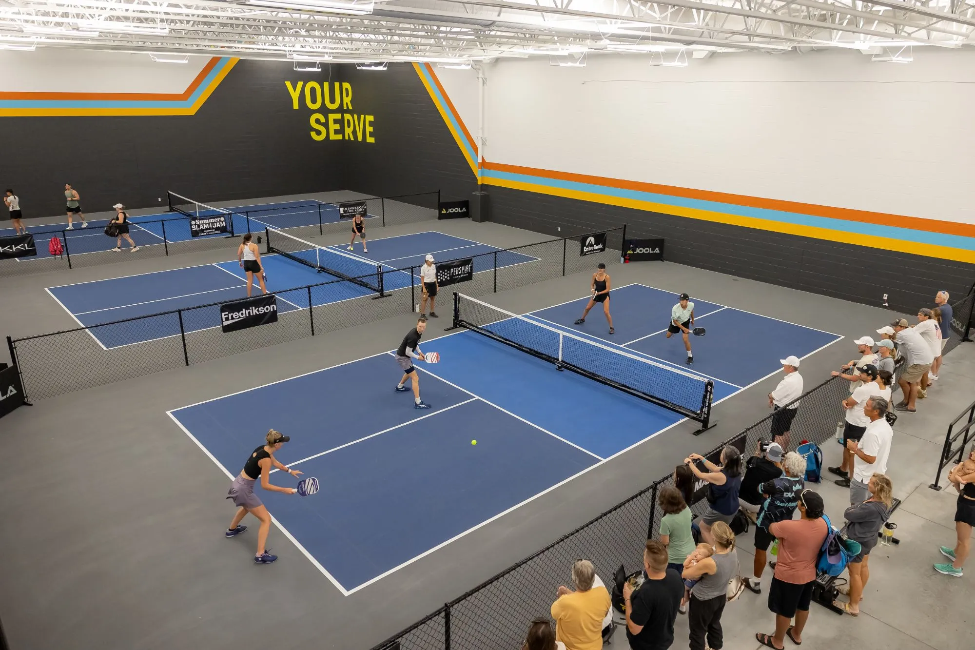 Elevated view of doubles pickleball on blue indoor courts beneath the Your Serve mural, with spectators along the rail