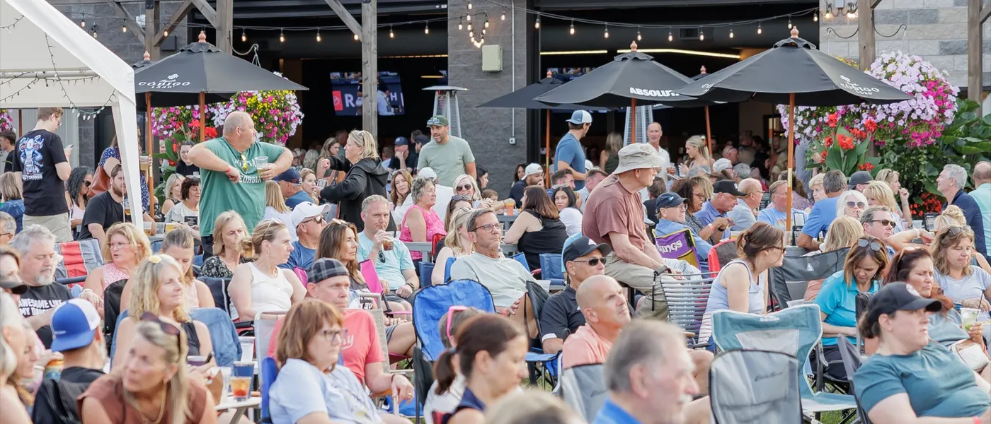 The crowd in camp chairs on the lawn facing the building, pergola string lights and hanging flower baskets behind