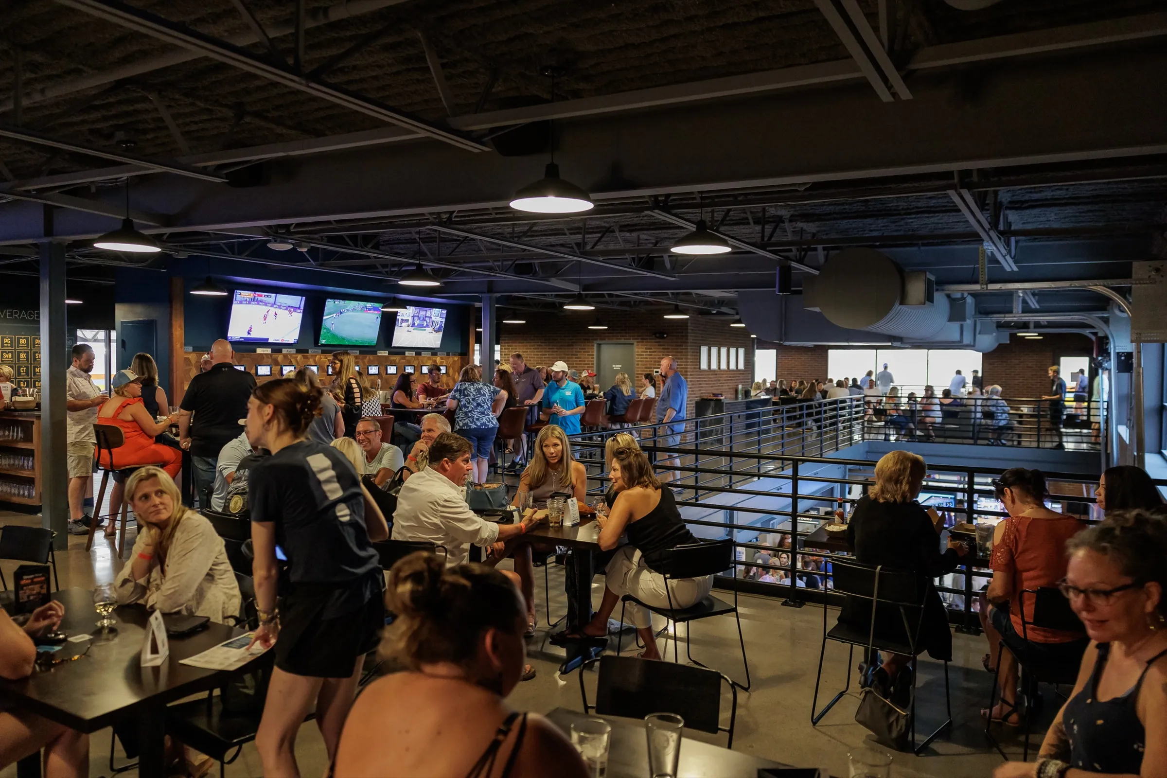 The mezzanine during an event, guests at high-top tables with the bar and TVs behind and the courts below the railing
